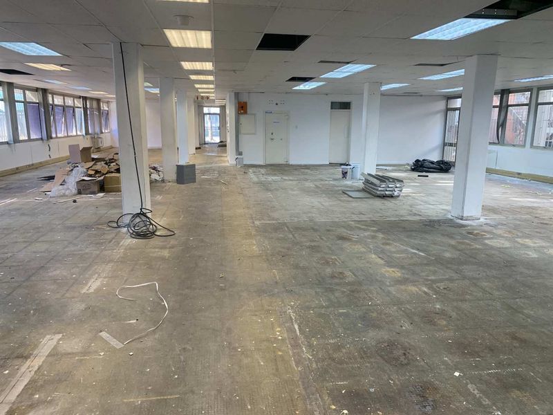A large, open-plan industrial office space featuring structural columns, suspended ceilings, and expansive windows, currently in need of refurbishment and flooring replacement.