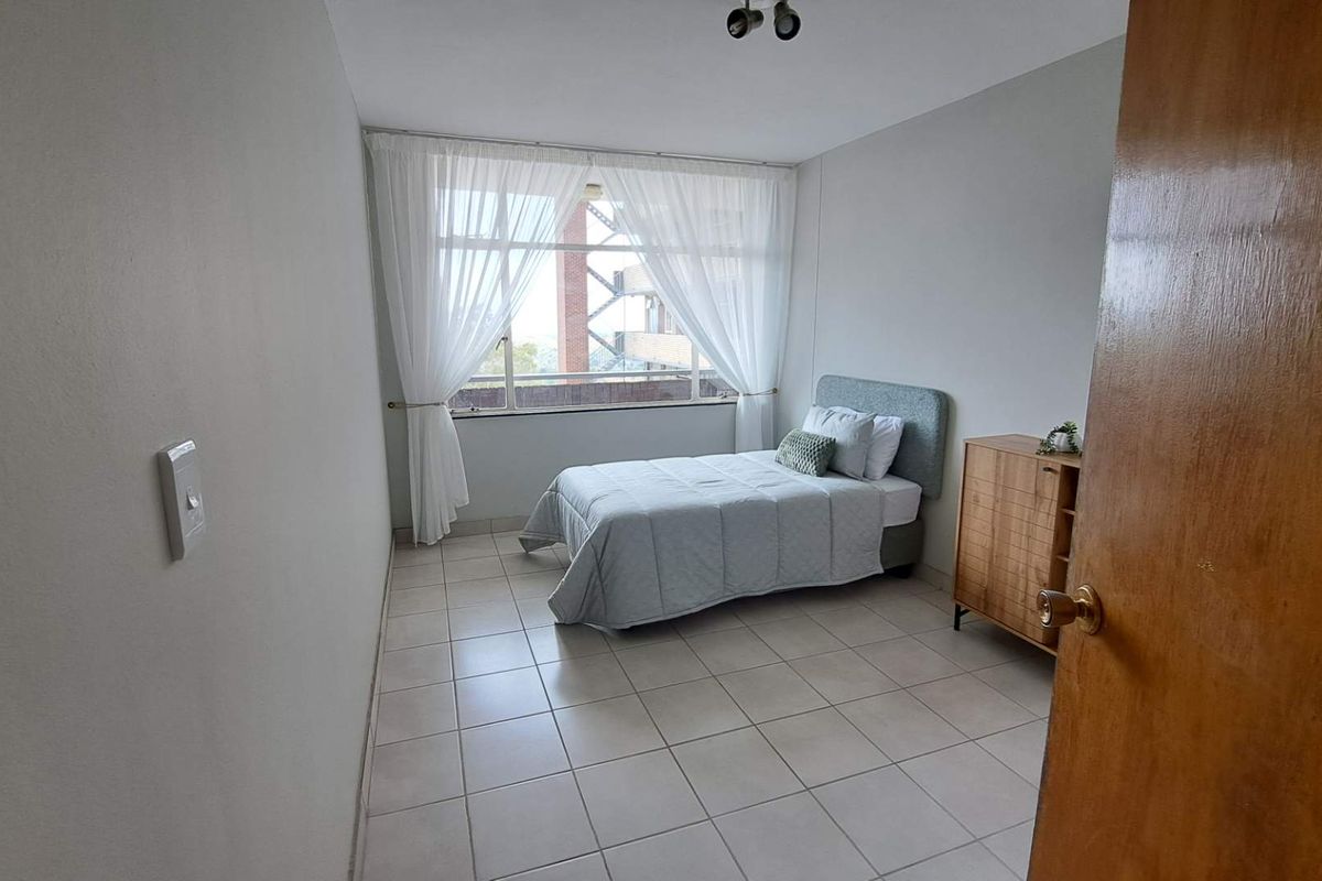 A functional bedroom featuring neutral tiled flooring, large window with natural light, and a standard door entrance.