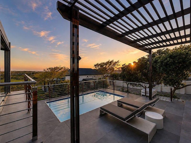 An elevated outdoor pool area featuring a timber pergola, modern tiled patio, and stainless steel balustrades, offering scenic sunset views.