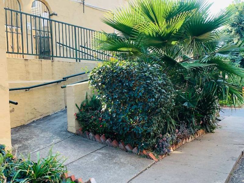 Exterior view of the townhouse entrance featuring a concrete ramp with handrails, metal security fencing, and established garden beds with brick edging.