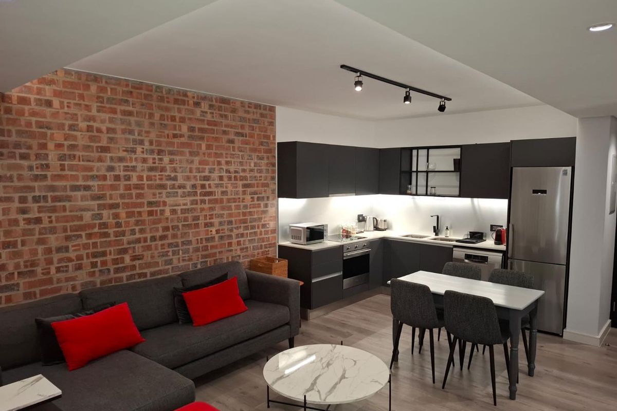 Modern open-plan living area featuring a striking exposed brick wall, contemporary dark cabinetry, integrated kitchen appliances, and stylish track lighting.