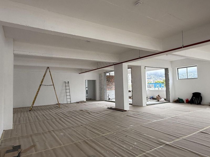 Spacious open-plan office area in shell condition, featuring exposed structural beams, fire sprinkler piping, and large windows providing natural light.