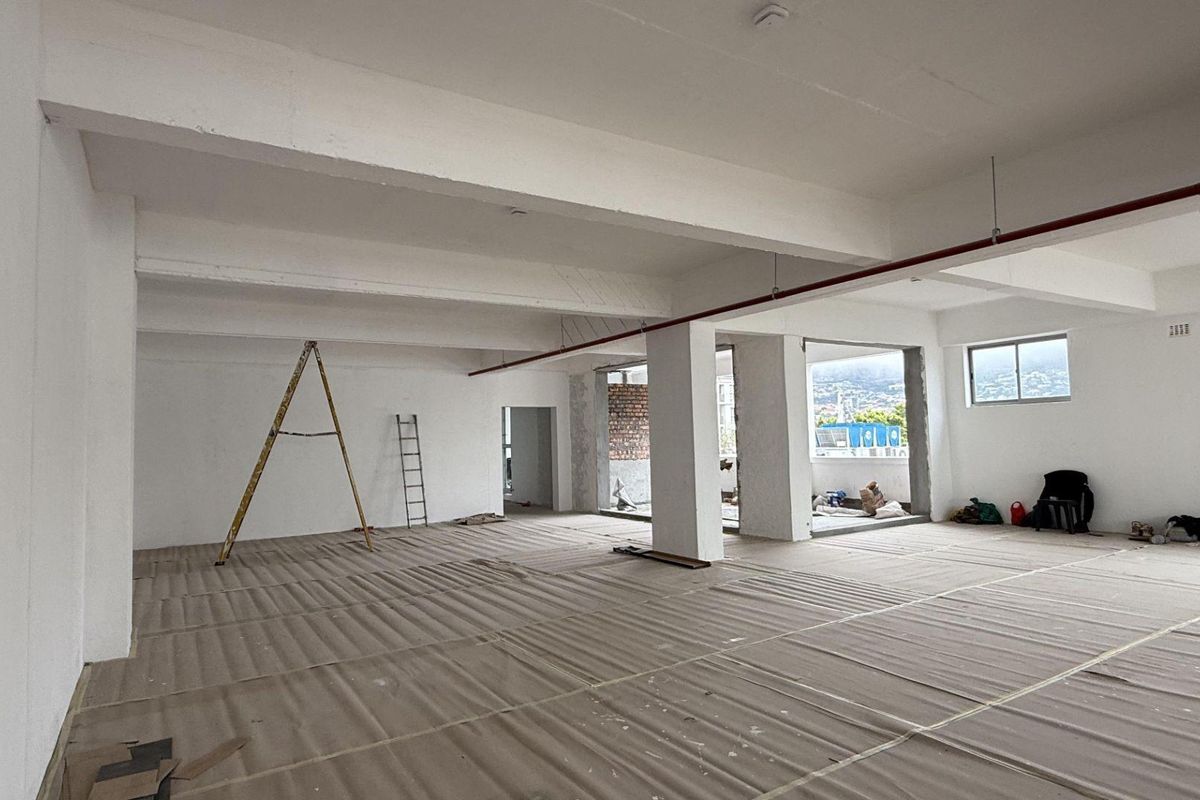 Spacious open-plan office area in shell condition, featuring exposed structural beams, fire sprinkler piping, and large windows providing natural light.