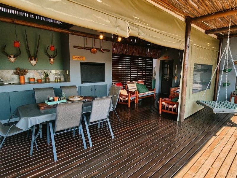 A spacious covered patio featuring timber decking, a thatched roof, built-in braai, and a kitchenette area, perfect for outdoor entertaining.