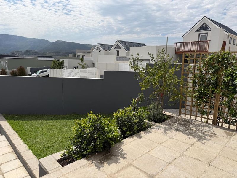A well-maintained rear garden featuring a paved patio area, a modern retaining wall, and a timber trellis, set against a backdrop of mountain views.