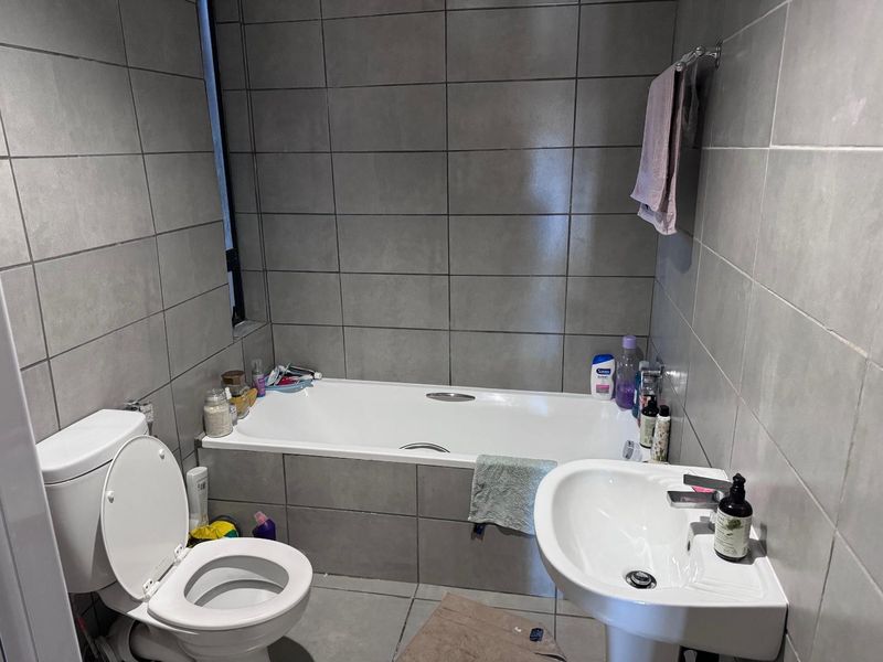 A functional bathroom featuring grey wall and floor tiles, a standard bathtub, a pedestal basin, and a toilet.