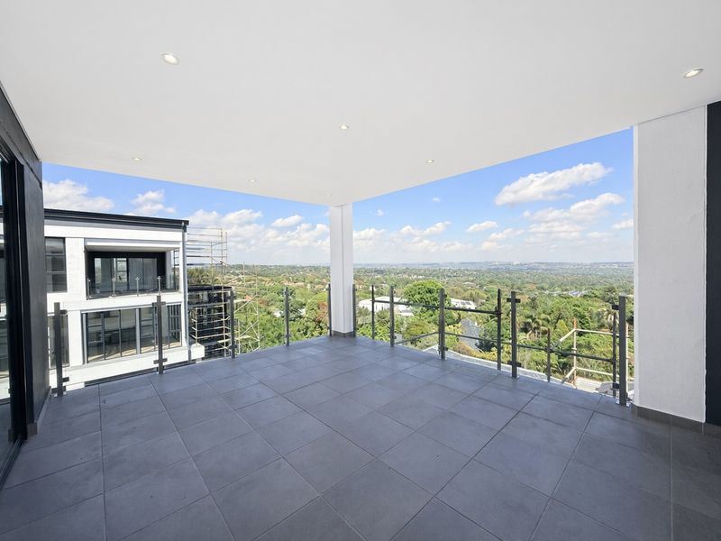 A spacious, modern covered balcony featuring tiled flooring, a sleek glass balustrade, and expansive elevated views of the surrounding landscape.