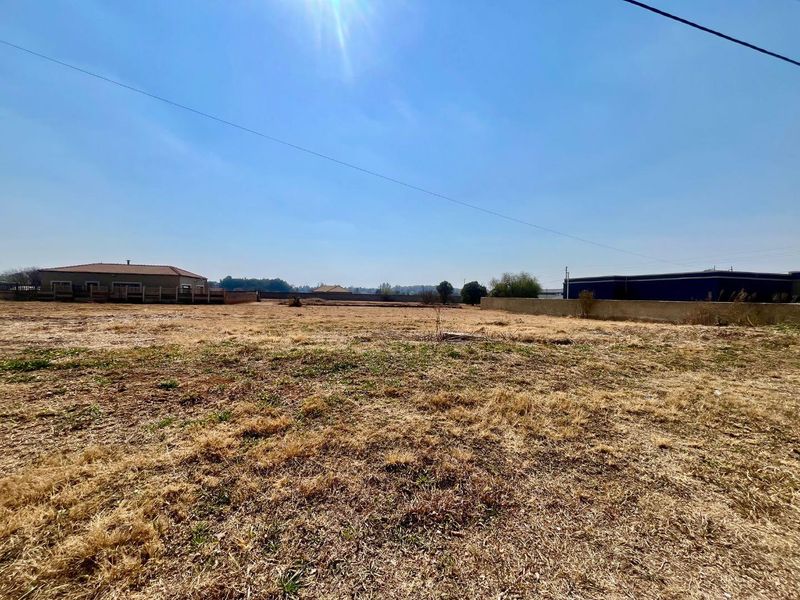 A wide-angle view of a vacant residential plot featuring dry grass, perimeter boundary walls, and surrounding neighborhood structures.