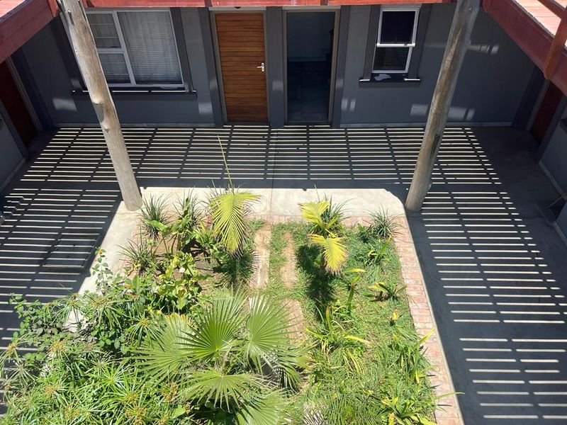 A view of a central courtyard featuring a concrete patio, landscaped garden beds, and a timber pergola structure providing shade to the building entrance.