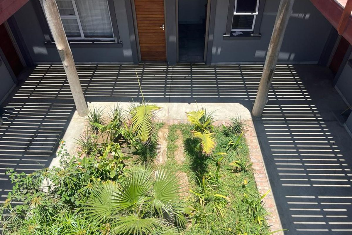 A view of a central courtyard featuring a concrete patio, landscaped garden beds, and a timber pergola structure providing shade to the building entrance.