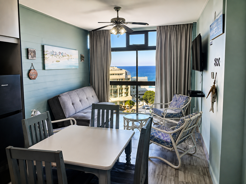 Open-plan living and dining area featuring light blue panelled walls, laminate flooring, and a large sliding door offering ocean views.