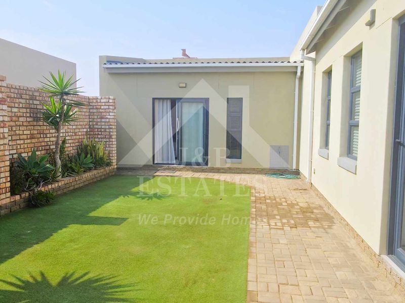 A well-maintained rear courtyard featuring a paved walkway, artificial lawn, and a brick boundary wall with a raised garden bed.