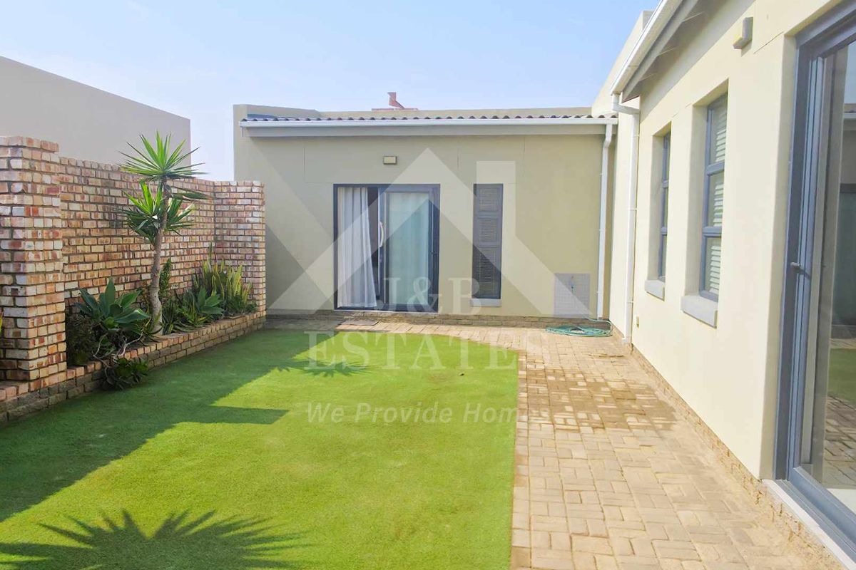 A well-maintained rear courtyard featuring a paved walkway, artificial lawn, and a brick boundary wall with a raised garden bed.