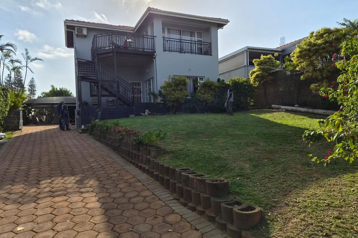 Rear view of the property featuring a paved driveway, landscaped lawn with retaining wall, and an elevated balcony with external staircase access.