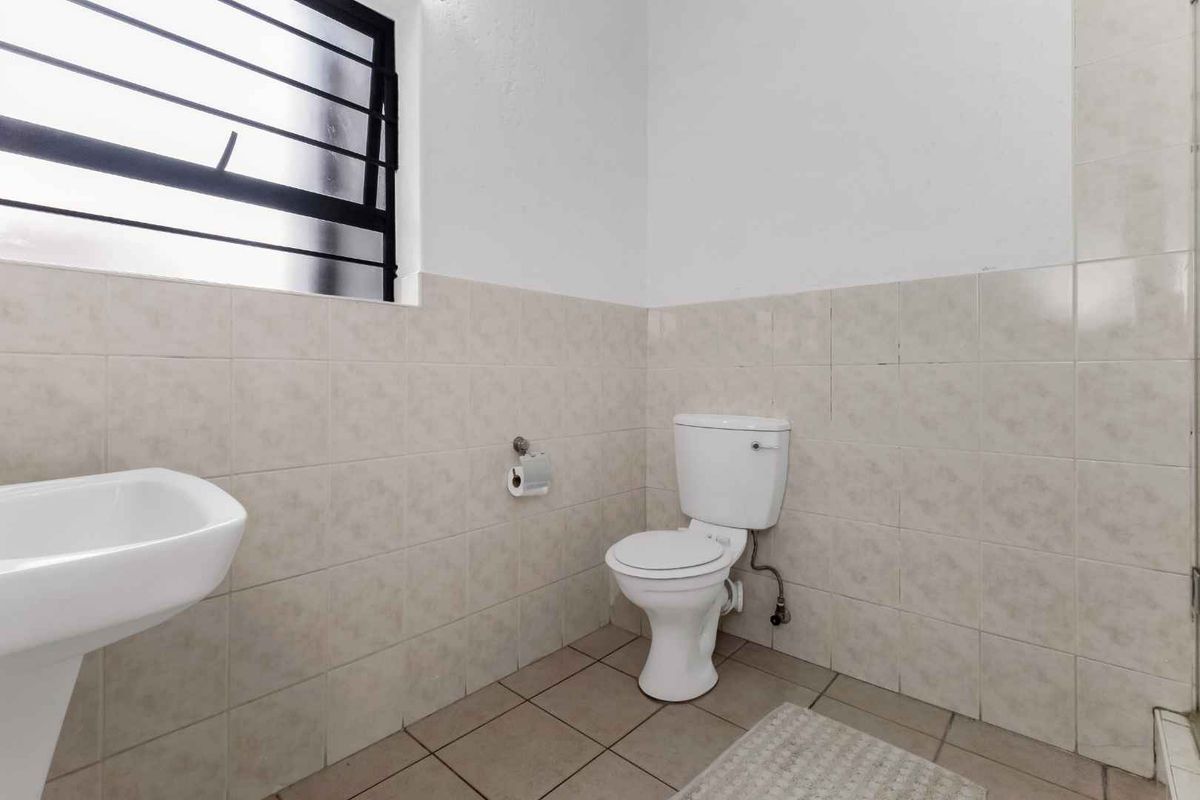 A functional bathroom featuring a pedestal sink, a standard toilet, and neutral wall tiling with a security-barred window.