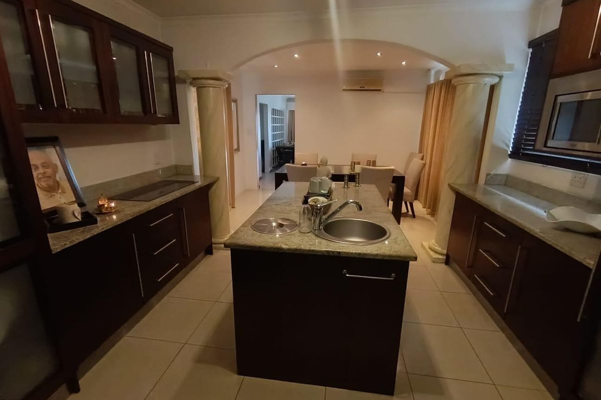 A well-appointed kitchen featuring dark wood cabinetry, granite countertops, a central island with a sink, and decorative columns leading to the dining area.