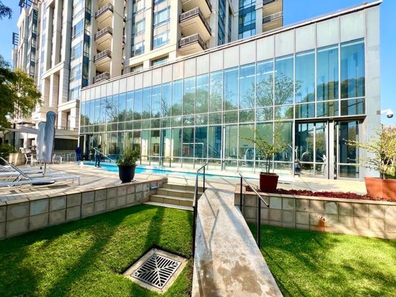 Outdoor pool area featuring a modern glass-enclosed facility, tiled retaining walls, and a concrete ramp access point within a landscaped apartment complex setting.
