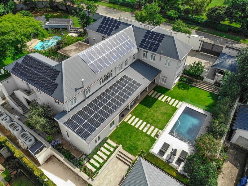 Aerial view of a well-maintained residential property featuring extensive solar panel installations, a private swimming pool, landscaped gardens, and secure boundary walls.