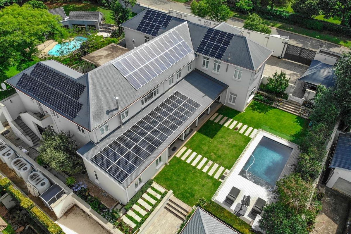 Aerial view of a well-maintained residential property featuring extensive solar panel installations, a private swimming pool, landscaped gardens, and secure boundary walls.