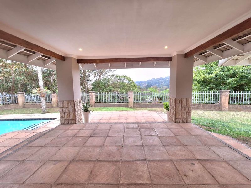 A spacious covered patio featuring stone-clad pillars and exposed timber beams, overlooking a private swimming pool and landscaped garden area.