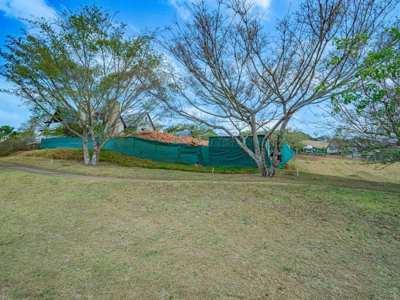 A view of the rear residential land featuring mature trees, a grassy lawn, and temporary construction fencing surrounding a building site.