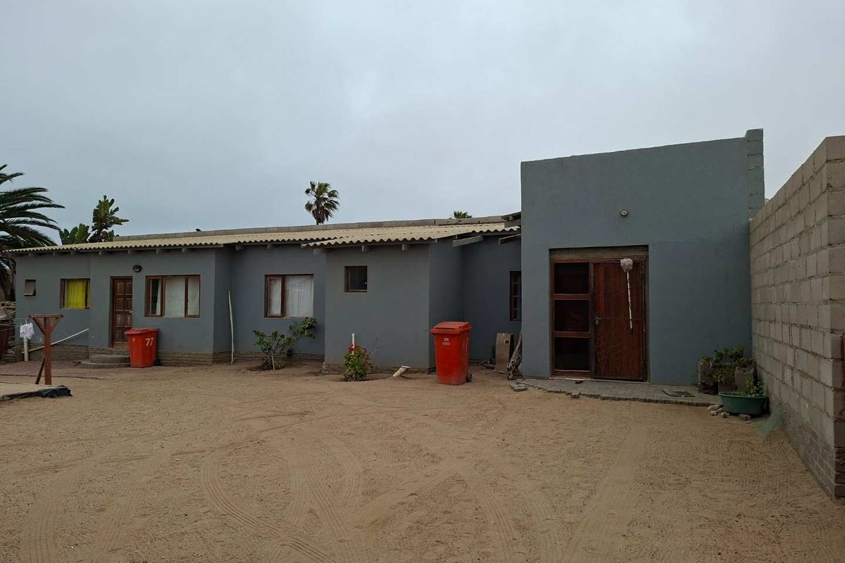 Exterior view of a single-story house featuring a grey rendered facade, wooden door, and an expansive unpaved sandy yard area.