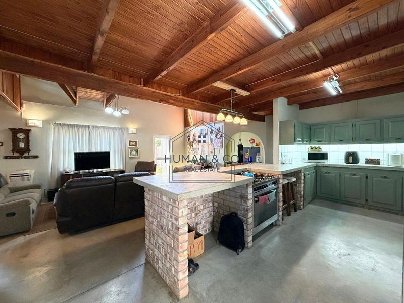 Spacious open-plan living and kitchen area featuring exposed timber beam ceilings, a brick kitchen island, gas stove, and durable concrete flooring.