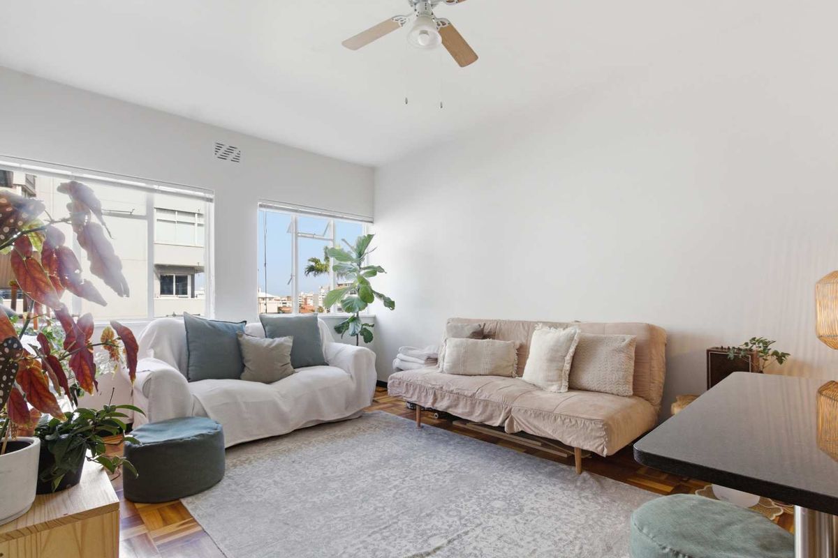 A bright living room featuring classic parquet flooring, a ceiling fan, and large windows providing natural light and an urban outlook.