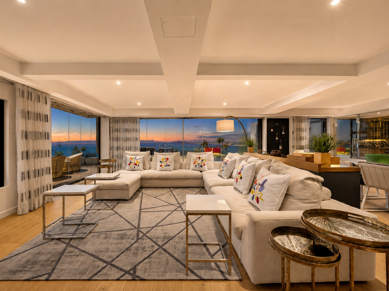 Spacious open-plan living area featuring hardwood floors, recessed lighting, and expansive floor-to-ceiling glass doors offering panoramic sunset views.