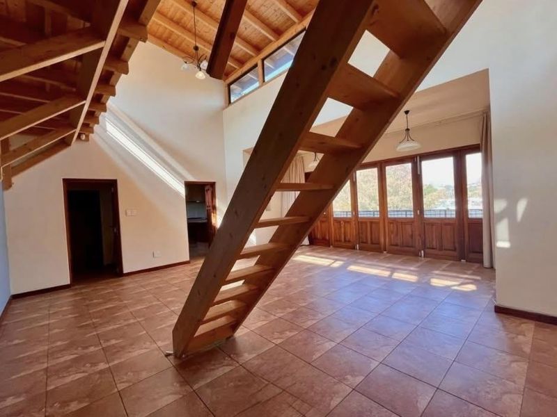 Spacious open-plan living area featuring high vaulted ceilings with exposed timber beams, tiled flooring, and large timber-framed glass doors leading to an outdoor space.