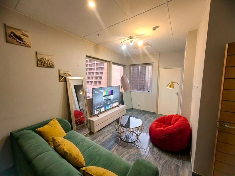 A compact living room featuring tiled flooring, a drop ceiling with integrated lighting, and large windows providing natural light and urban views.