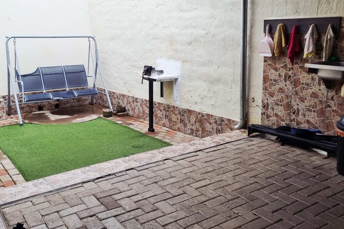 A paved rear courtyard area featuring a small artificial grass section, a wall-mounted utility sink, and tiled wall accents.