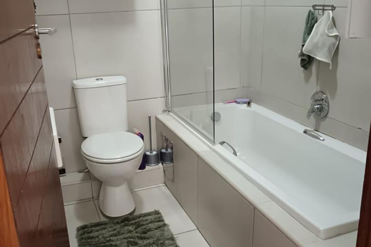 A functional bathroom featuring a bathtub with a glass shower screen, tiled walls and flooring, and a wall-mounted toilet.