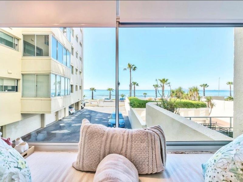 A bright interior view looking out through a large window towards the ocean, palm trees, and surrounding apartment complex.