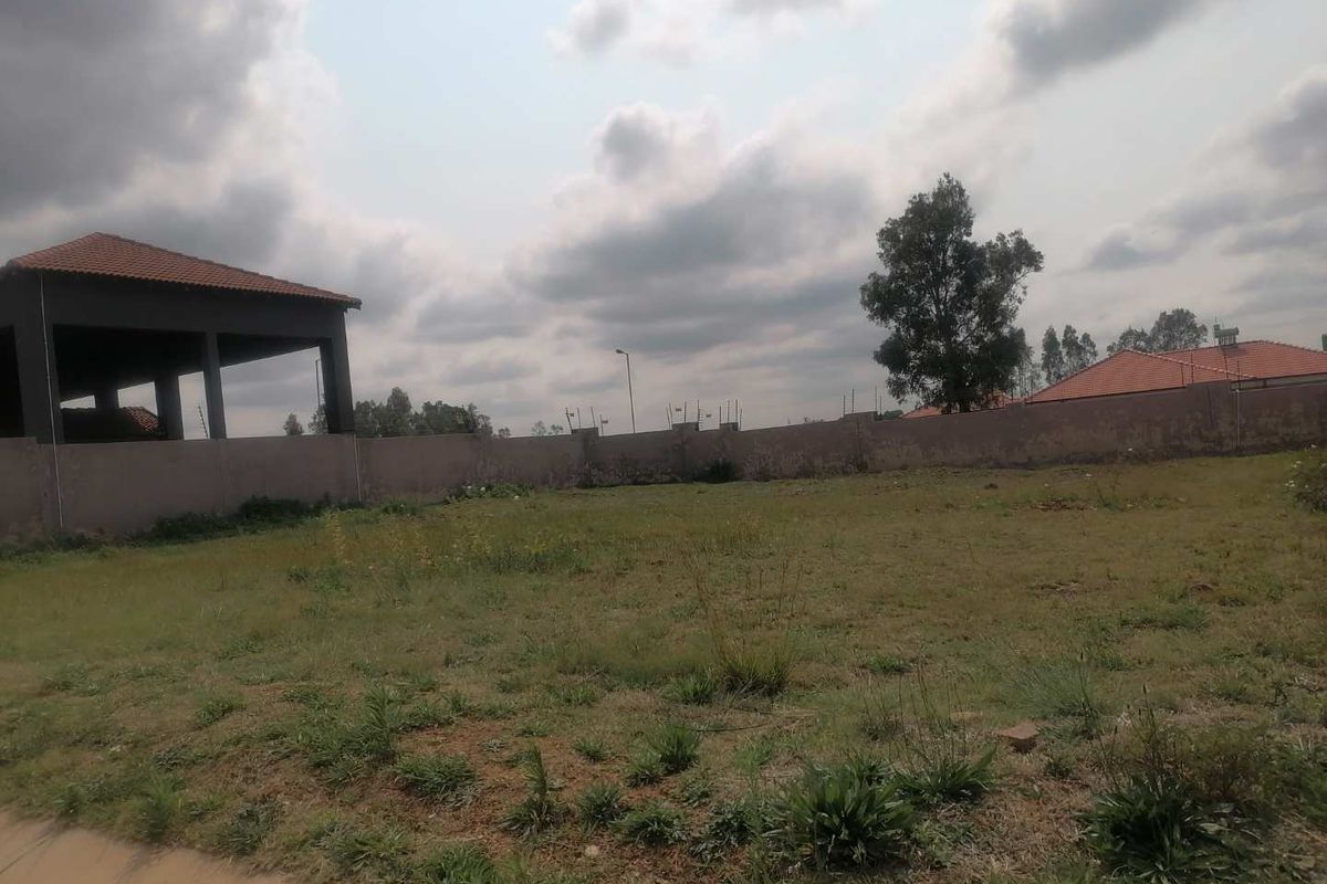 A vacant residential plot featuring a perimeter wall and an adjacent covered structure under a cloudy sky.