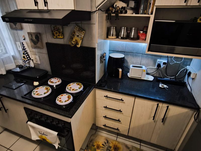 A functional kitchen featuring light wood-finish cabinetry, dark granite countertops, an integrated electric hob with extractor fan, and tiled flooring.