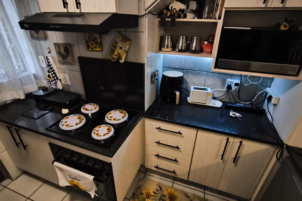 A functional kitchen featuring light wood-finish cabinetry, dark granite countertops, an integrated electric hob with extractor fan, and tiled flooring.
