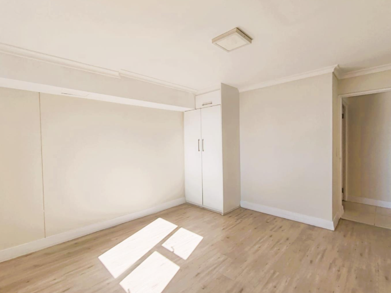 A bright, empty bedroom featuring light-toned laminate flooring, neutral walls, and a built-in wardrobe with clean finishes.