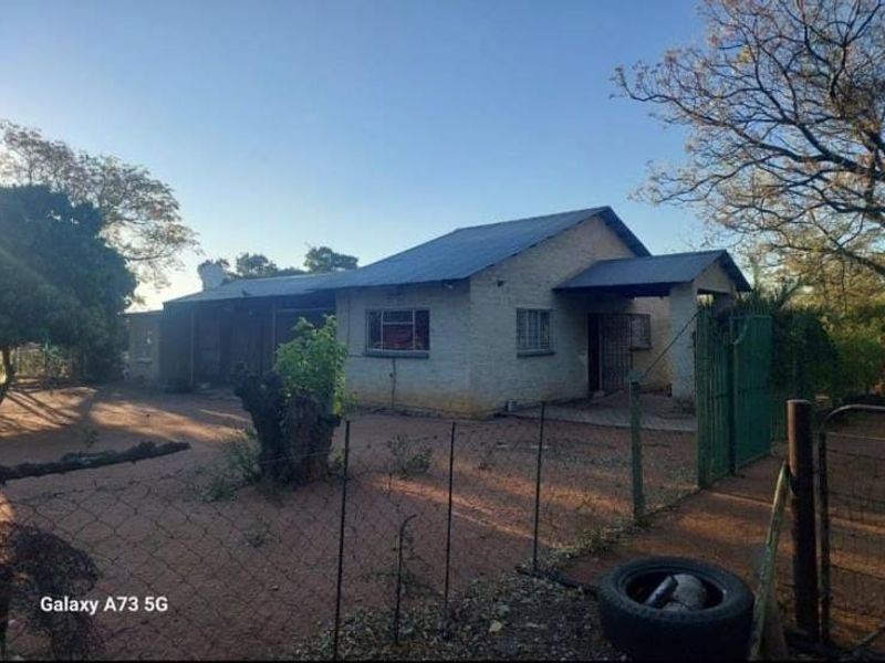 Front view of a single-story brick house with a metal roof, featuring security fencing and a gated entrance on a large plot.
