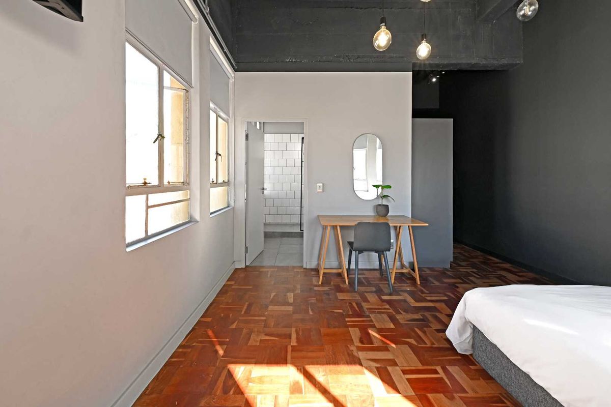 A spacious bedroom featuring classic parquet flooring, high exposed ceilings, and an en-suite bathroom with white subway tiling.