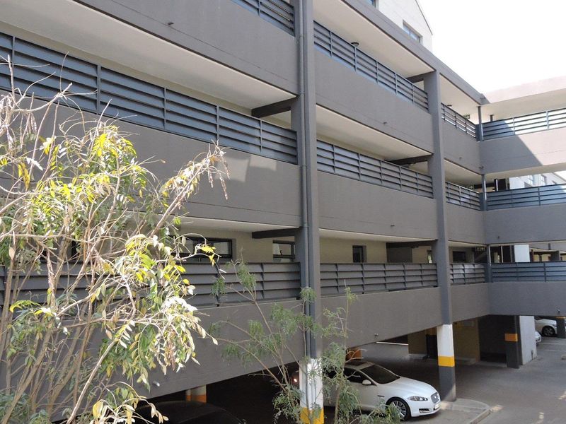 Exterior view of a multi-level apartment building featuring modern grey facade finishes, metal balcony railings, and a covered ground-level parking area.