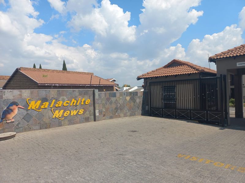 Front entrance view of Malachite Mews townhouse complex featuring a stone-clad boundary wall, secure metal gate, and paved visitor parking area.