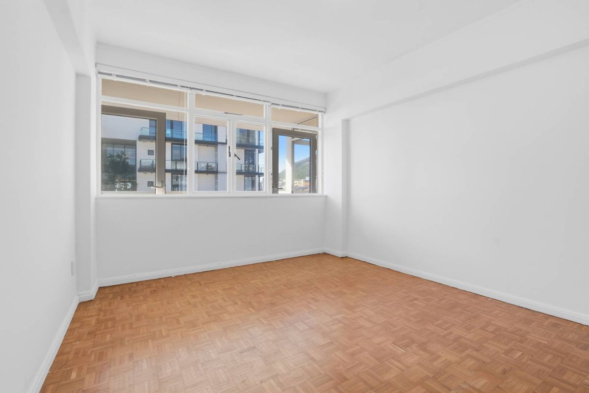 A bright, empty bedroom featuring classic parquet flooring and large windows that provide natural light and an urban outlook.