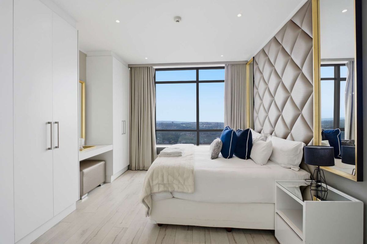 Modern penthouse bedroom featuring a large upholstered headboard, built-in floor-to-ceiling wardrobes, integrated vanity, and expansive city views through large windows.