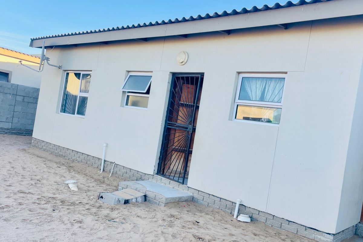 Exterior view of a single-story house featuring a security gate, aluminium windows, and an unfinished sandy yard area.