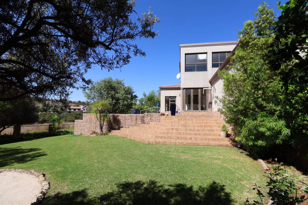 Rear view of the property featuring a spacious lawn, stone retaining wall, and a tiled patio area leading to the main residence.