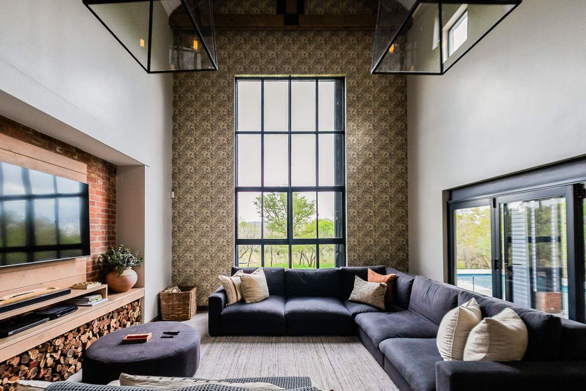 Spacious double-volume living room featuring a striking brick feature wall, large steel-framed windows, exposed timber beams, and seamless access to the outdoor area.