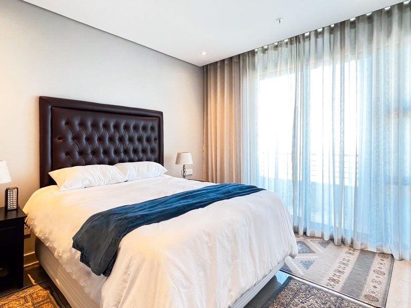 A well-maintained bedroom featuring neutral walls, recessed lighting, and large sliding glass doors with floor-to-ceiling curtains providing access to an outdoor area.