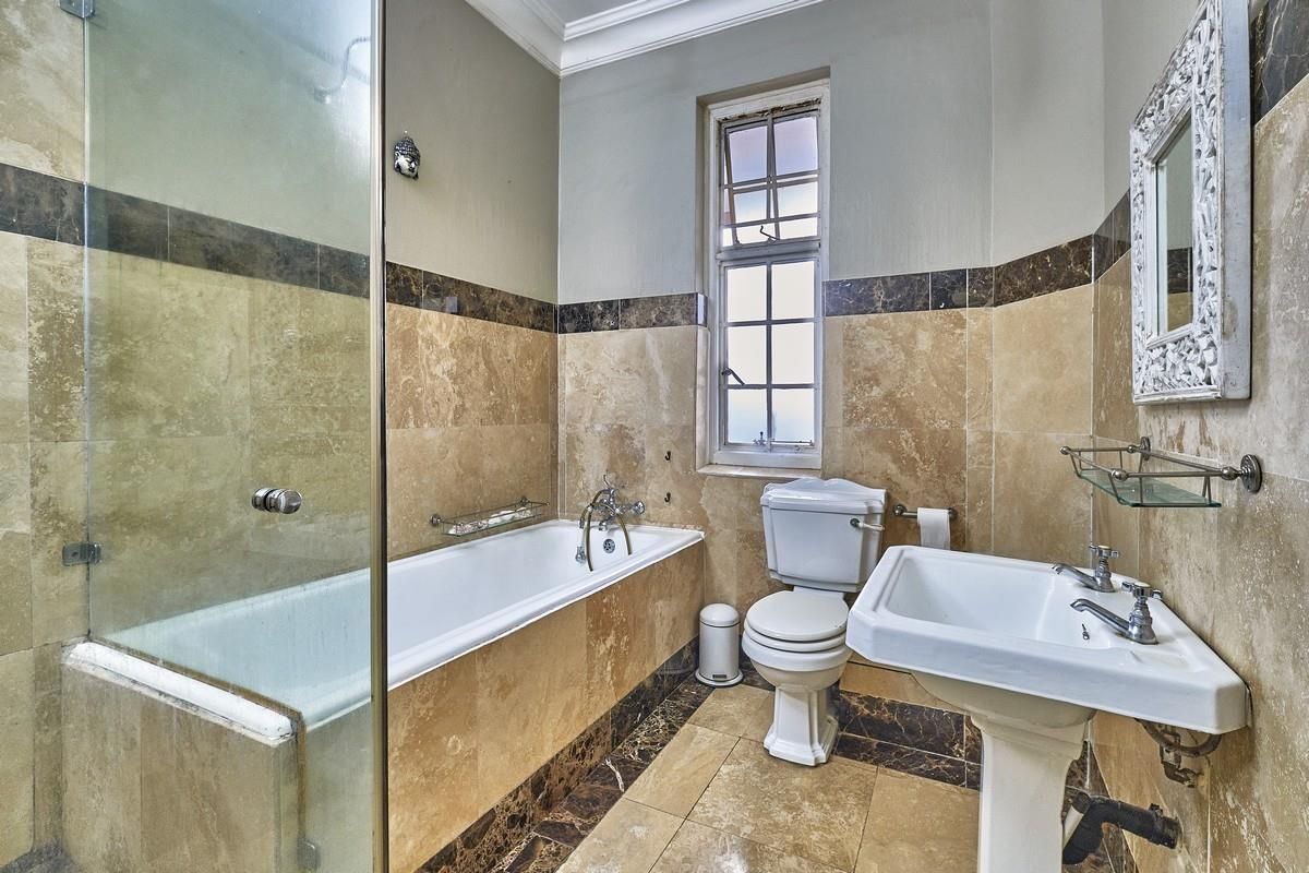 A classic bathroom featuring travertine wall and floor tiles, a built-in bathtub with glass shower screen, pedestal sink, and toilet.