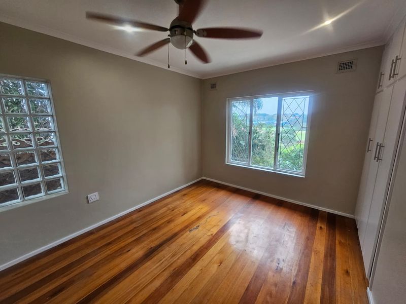 A bright, empty bedroom featuring polished timber flooring, built-in cupboards, a ceiling fan, and a unique glass block feature window.